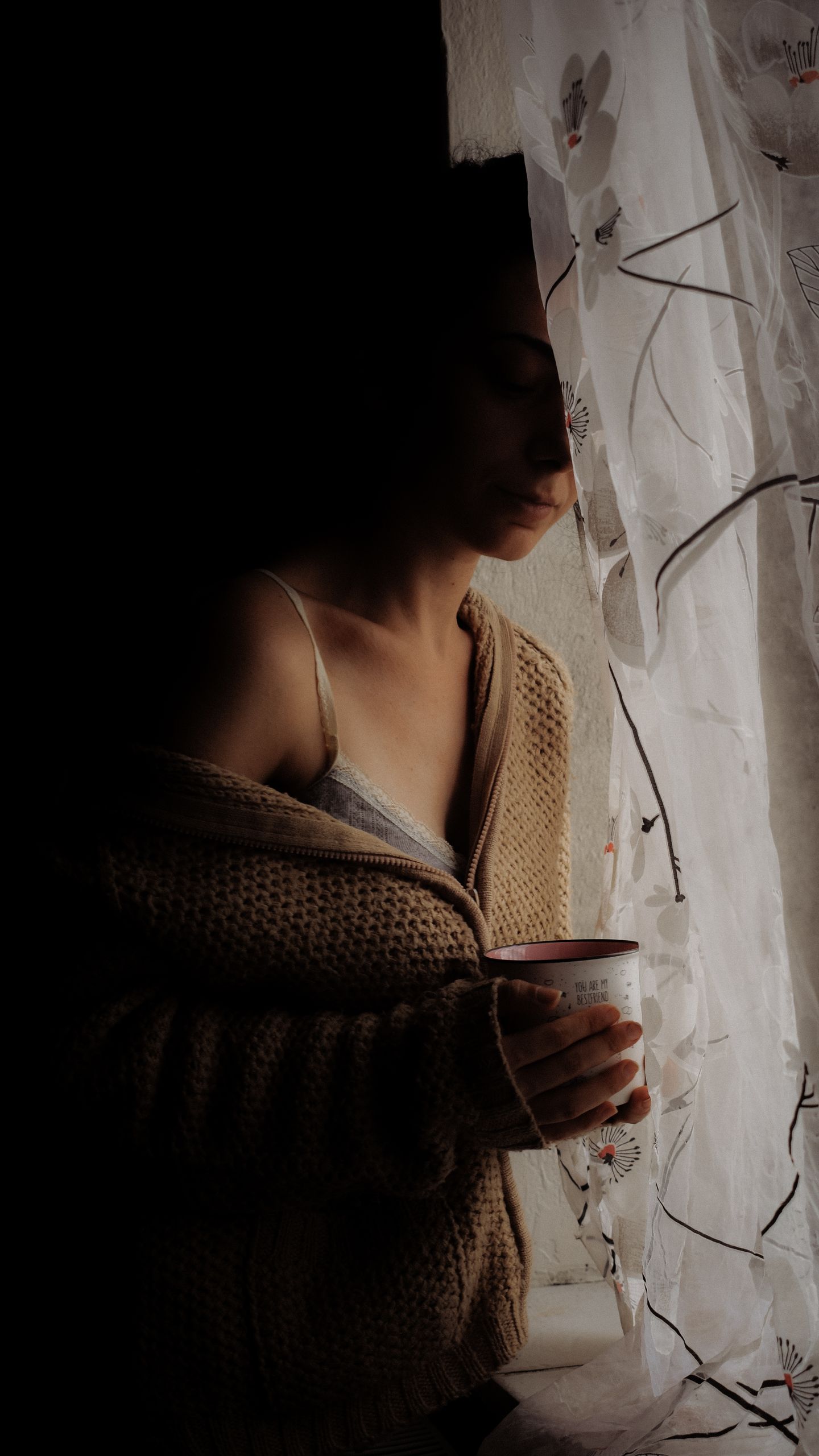 A woman holding a cup, standing by a window with soft lighting and shadows.